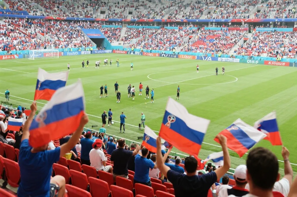 世界杯赛前训练场看台上，球迷举着旗帜等待球队亮相的热烈氛围